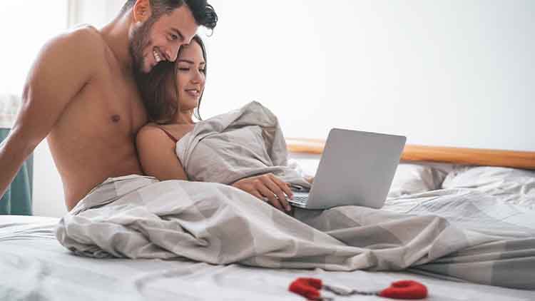 Happy couple watching on computer while lying on bed under blanket 